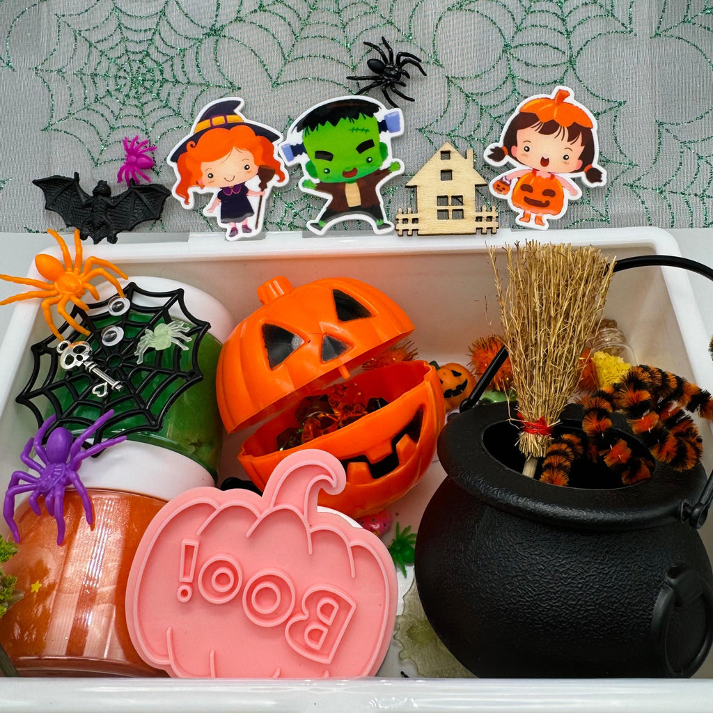 Halloween sensory play dough box with decorations including a jack-o’-lantern, black cauldron, spider web, witch, Frankenstein, pumpkin girl cutouts, plastic spiders, bats, and ‘Boo’ pumpkin mold for kids.