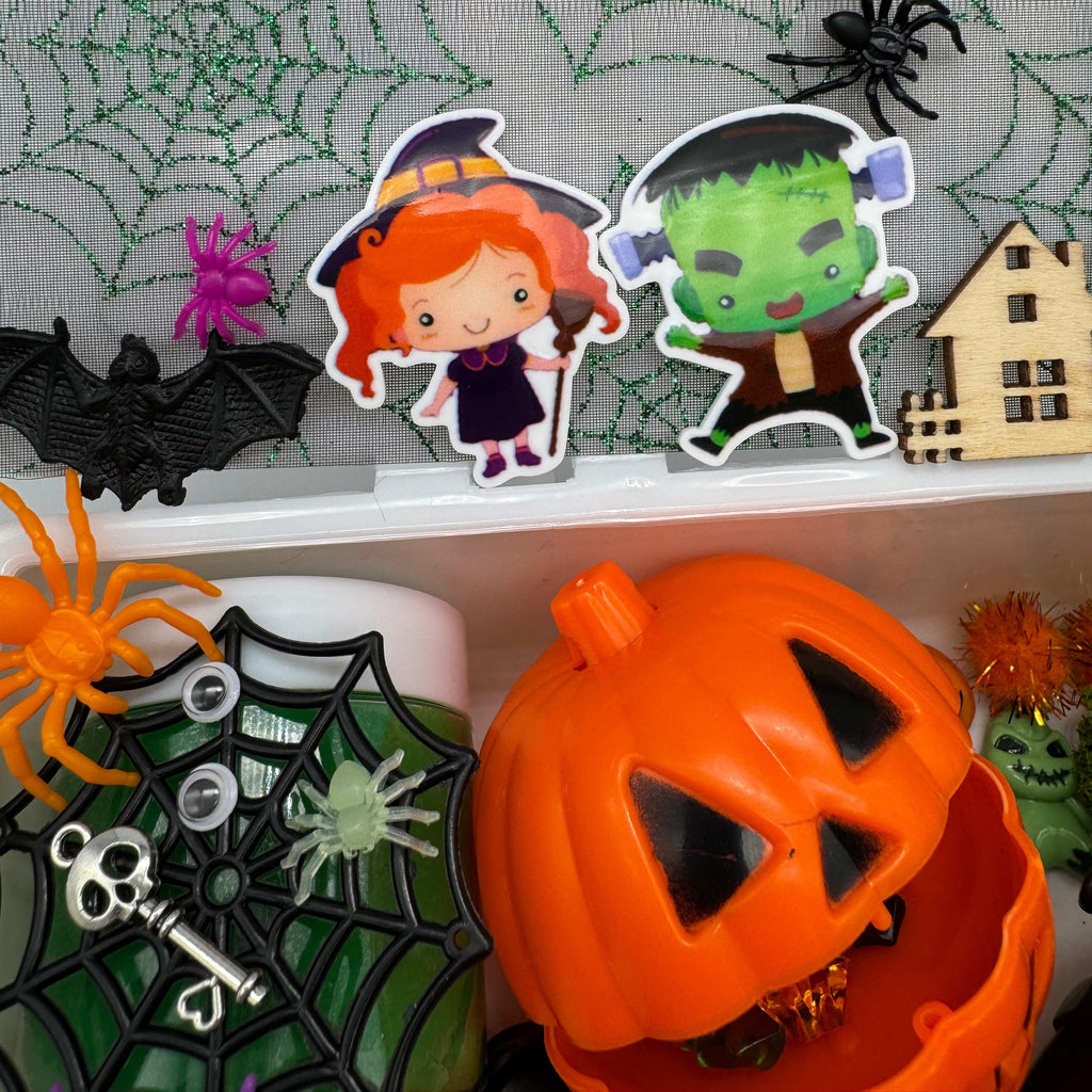 Halloween-themed sensory bo with witch and Frankenstein cutouts, plastic spiders, bat, jack-o’-lantern container, spider web decoration, skeleton key, and wooden haunted house prop